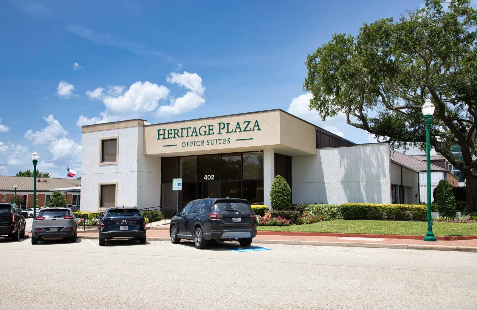 Heritage Plaza main entrance at 402 Simonton Street — the Office Suites signage above the glass doors, with parking and a Texas flag under a blue sky