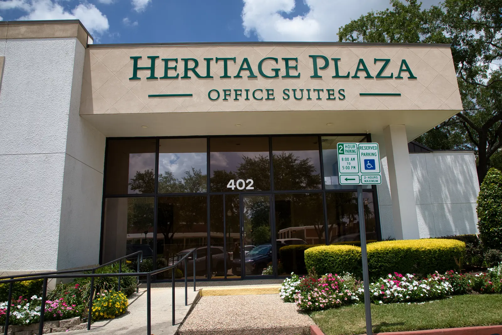 Close-up of the Heritage Plaza entrance and glass doors marked 402, framed by flower beds and clipped hedges